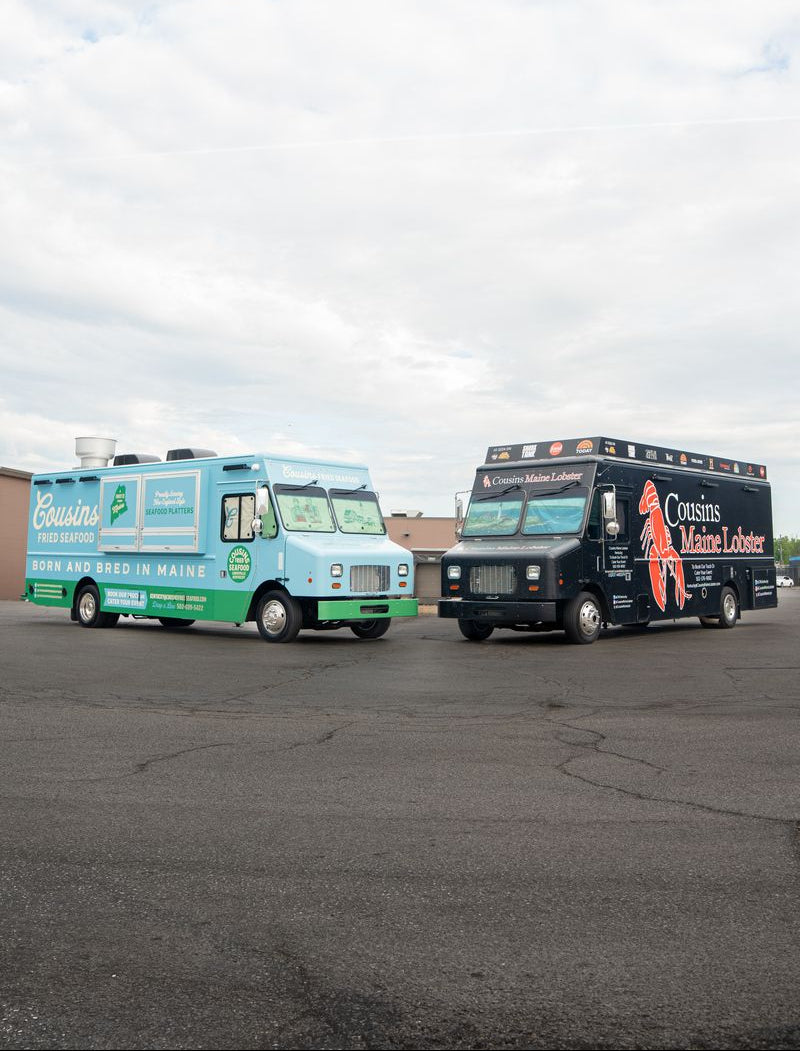 Two trucks, one blue and one black with branding, parked on a paved surface under a cloudy sky.
